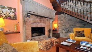 A cozy living room with a brick fireplace, a yellow armchair, a wooden coffee table, and a staircase with a wrought iron railing.