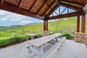 Uma área de piquenique coberta com bancos de madeira e um muro de pedra, com vista para uma paisagem verde com árvores e colinas.