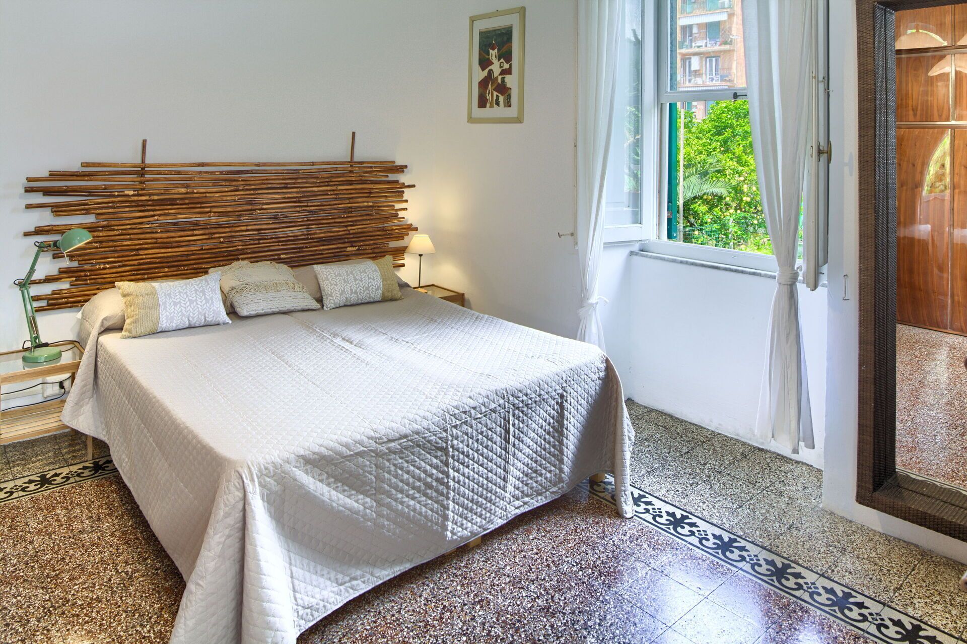 Una camera da letto con un letto, una testiera realizzata con listelli di bambù, una finestra che si affaccia sul verde e una porta che dà su un balcone.