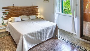A bedroom with a bed, a headboard made of bamboo sticks, a window with a view of greenery, and a door to a balcony. - Spacious apartment with garden, near the beach (Levanto)