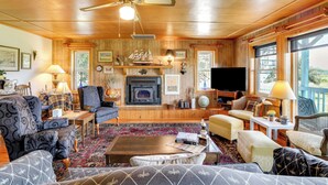 A cozy living room with wooden walls, a fireplace, and a large window. - Spacious Riverfront Cottage w/ Dock in Green Pond! (Green Pond)