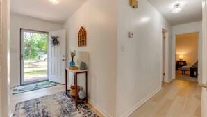A hallway with a wooden floor, a door leading outside, a small table with decorative items, and a wall-mounted light fixture. - Walk to Lake Munson: Tree-Filled Tallahassee Gem! (Tallahassee)