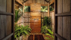 Un recinto de ducha de madera con techo de paja, rodeado de plantas tropicales y una terraza de madera.