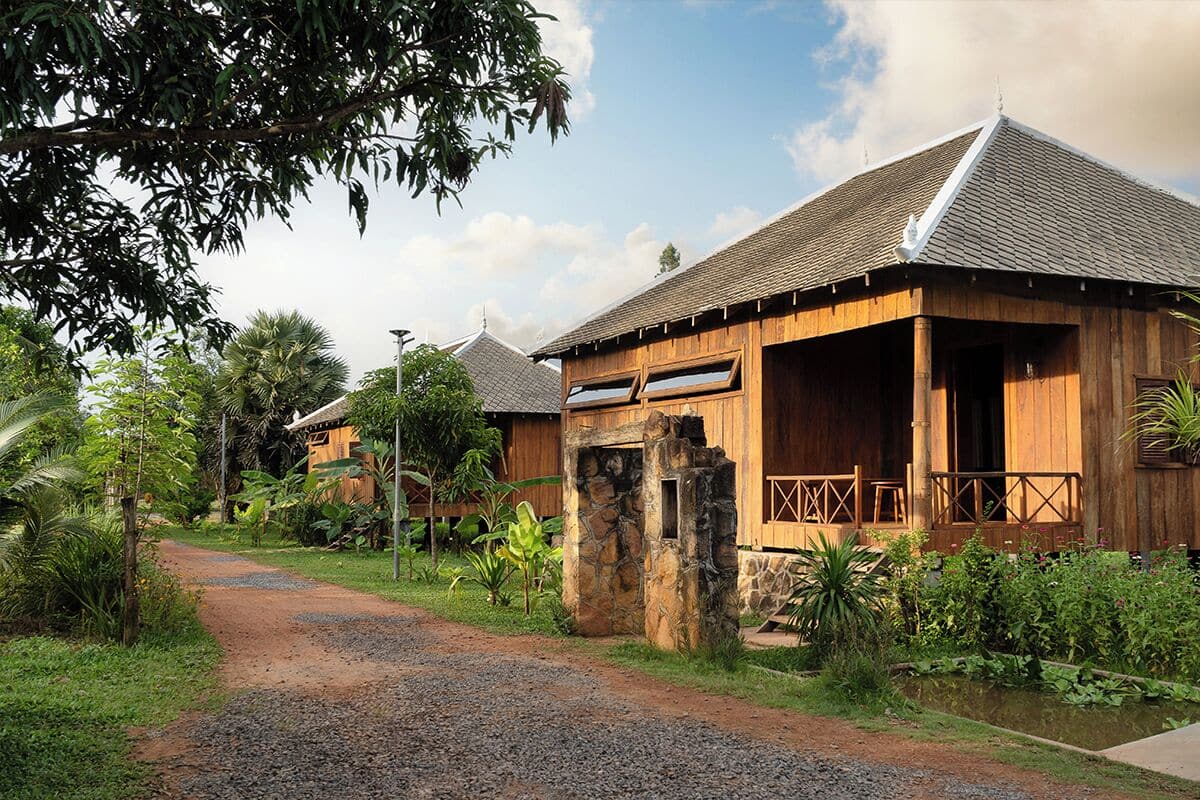 A rustic wooden building with a stone entrance, surrounded by lush greenery and a gravel path.