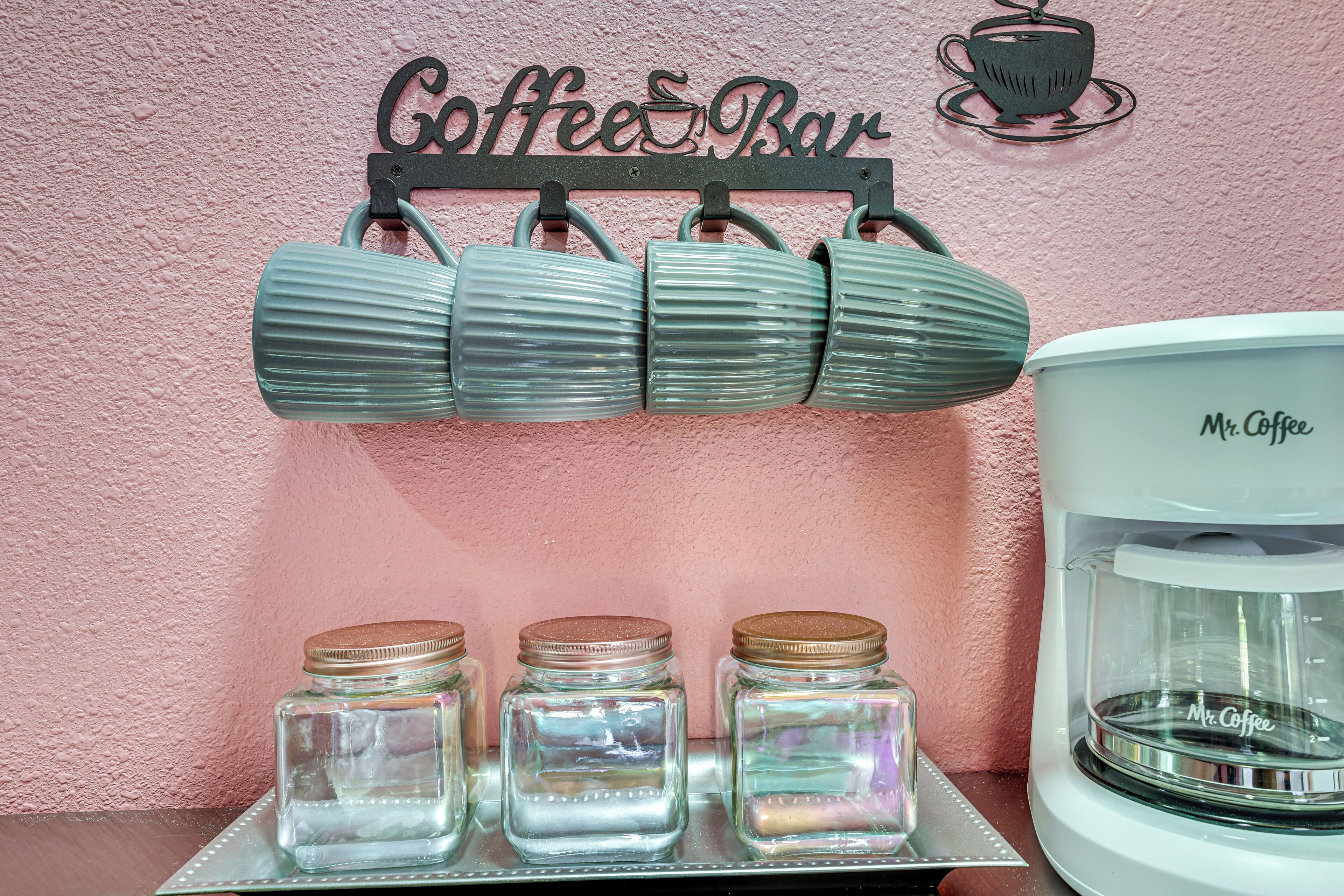 Un coin café avec une machine à café, des tasses et des bocaux, le tout disposé sur une table devant un mur rose.