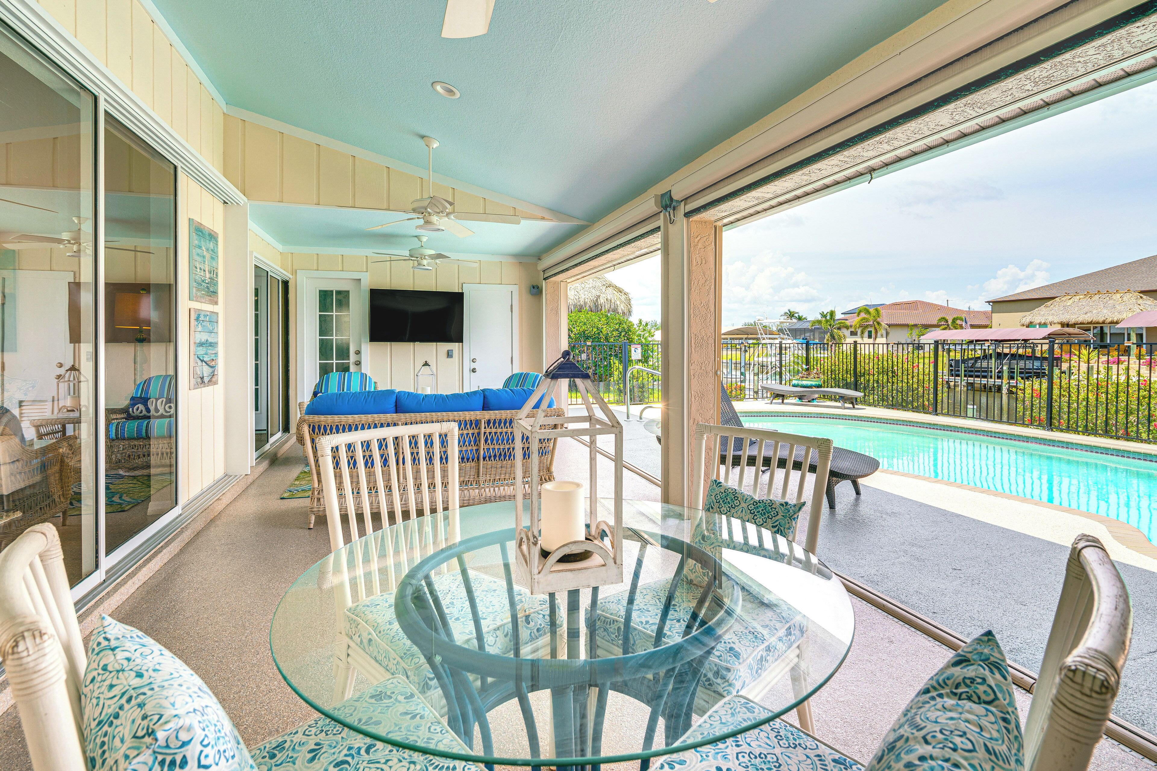 Ein Tisch mit Glasplatte, Stühle und Kissen, ein Deckenventilator und Blick auf einen Pool und einen Außenbereich.
