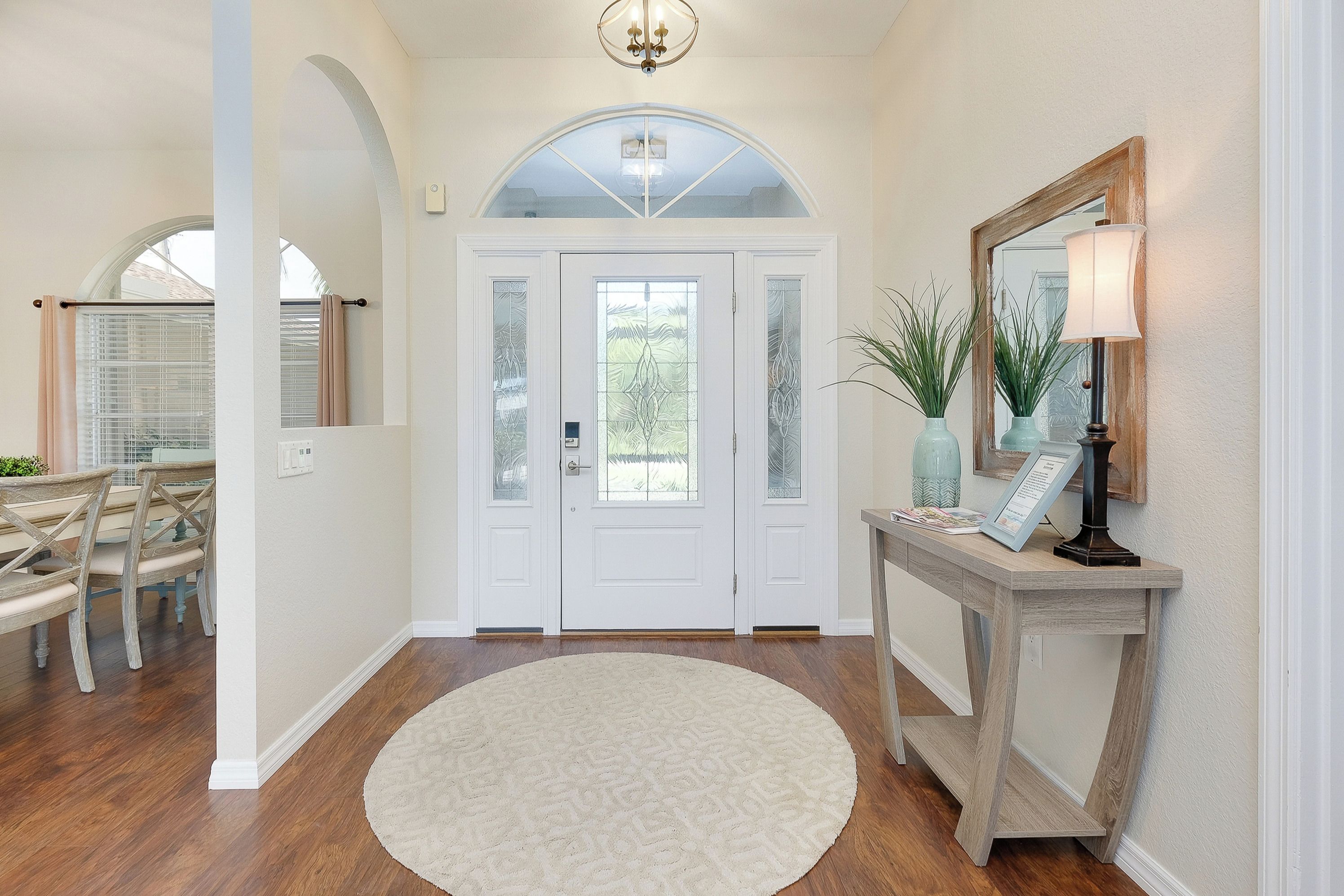 A foyer with a white door, a round rug, a console table with a mirror, and a dining area with wooden chairs.