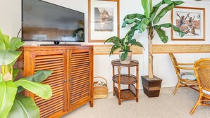 A living room with a television, framed artwork, a large potted plant, wicker furniture, and a woven basket. - Oceanfront Condo: Stunning Sunrise & Ocean Views (Hauula)