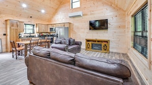 Interior de una cabaña de troncos con comedor, cocina, sala de estar con sofá de cuero y televisor en la pared.