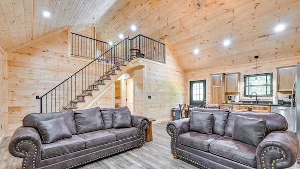 Interior espacioso de una cabaña de troncos con techo alto, una escalera, dos sofás de cuero, una zona de cocina y una mesa de comedor.