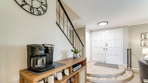 Un intérieur de maison avec une machine à café posée sur une console en bois, un escalier, une porte blanche et un fauteuil gris.