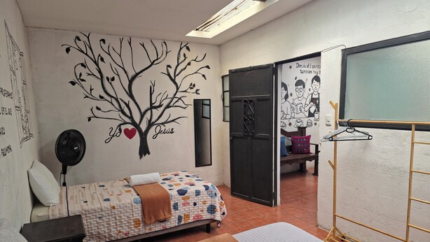 Un dormitorio con cama, mesita de noche, ventilador de pared, toalla, banco y tendedero.