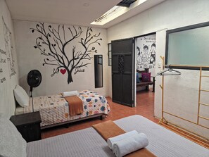 Une chambre à coucher comprenant un lit, une table de chevet, un ventilateur fixé au mur, une serviette, un banc et un étendoir.