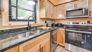 Une cuisine avec des meubles en bois, un plan de travail en granit, un évier en acier inoxydable, un four à micro-ondes, une cuisinière et une fenêtre avec des stores.