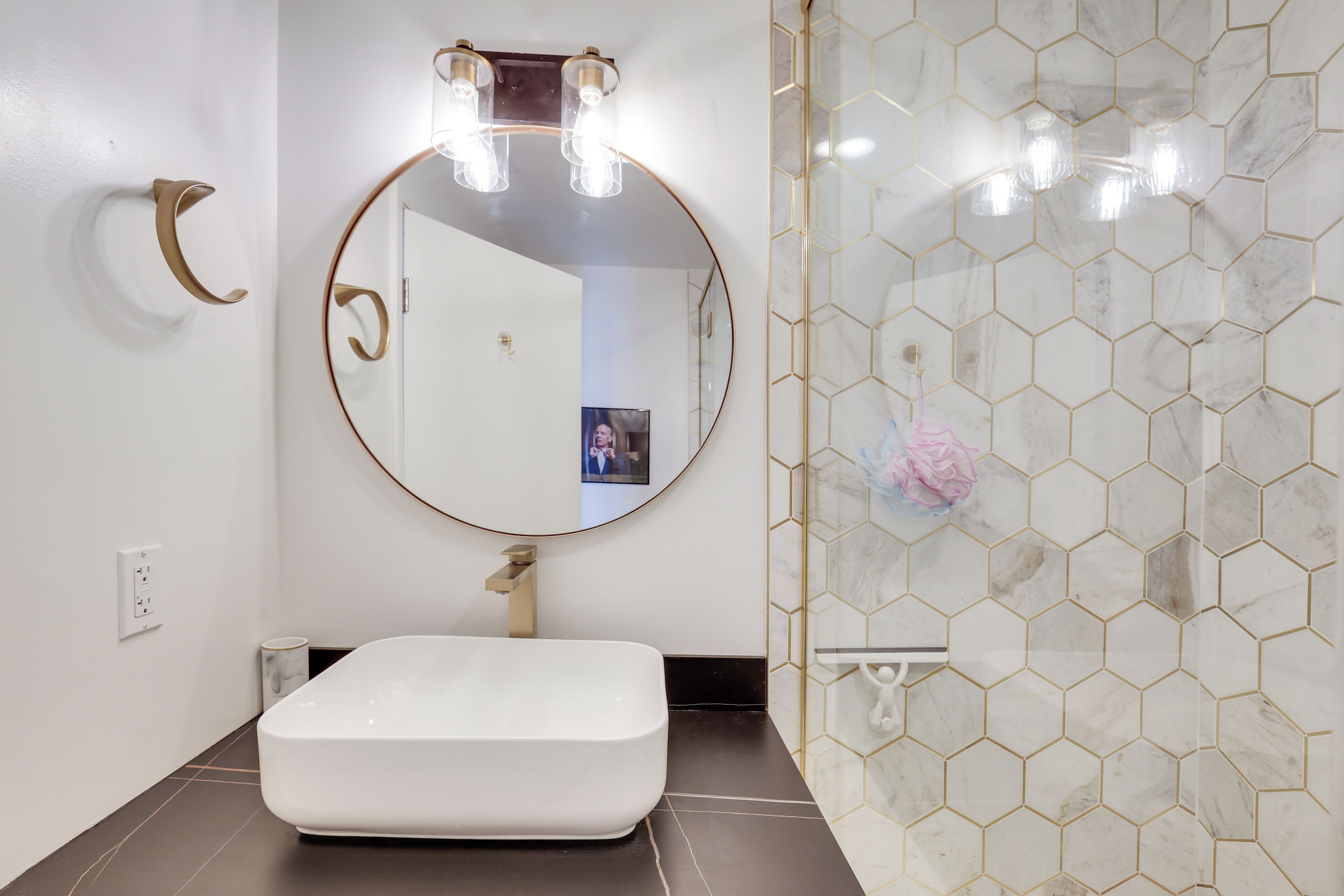 A modern bathroom with a white sink, a round mirror with three lights, and a hexagonal tile shower wall.