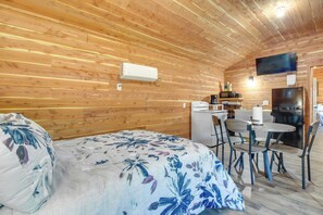Une pièce aux murs en bois, un lit avec une literie à motifs floraux, une table à manger avec des chaises, un réfrigérateur, un four à micro-ondes et un téléviseur à écran plat fixé au mur.