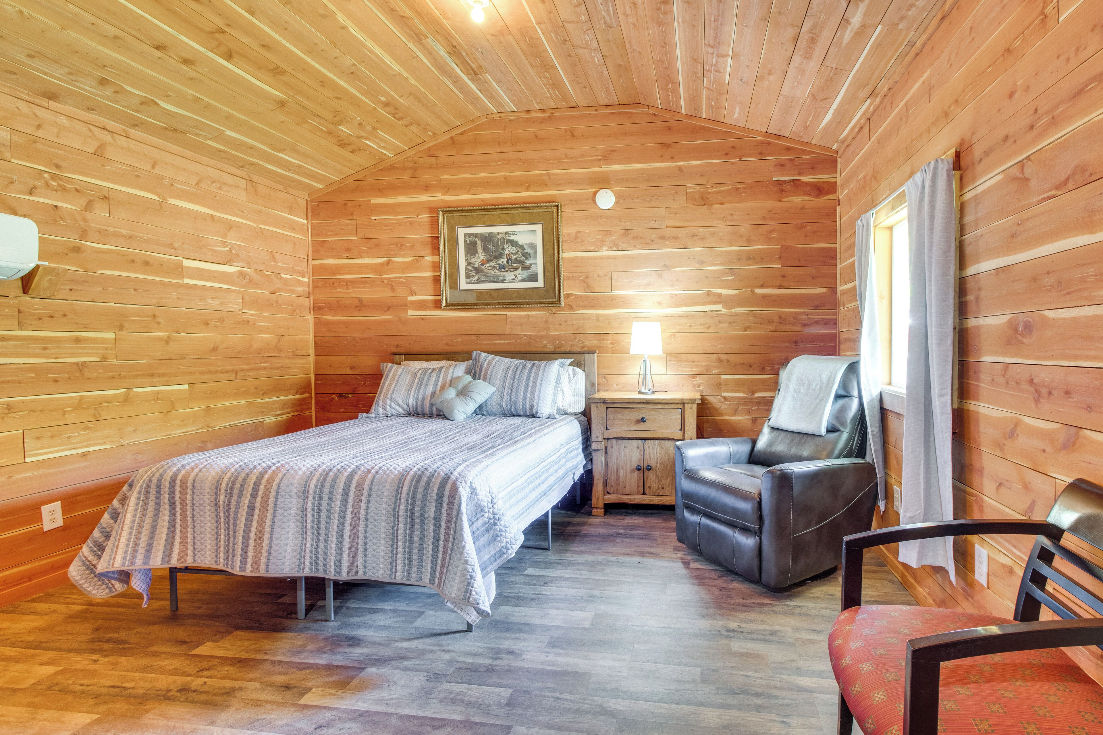 Een knus slaapkamer in een houten blokhut, met een bed, nachtkastje, stoel en een ingelijste foto aan de muur.