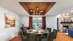 Un espace repas avec une table ronde dressée pour six personnes, entourée de chaises à revêtement vert. Un plafond en bois surmonté d’un luminaire moderne se trouve au-dessus.