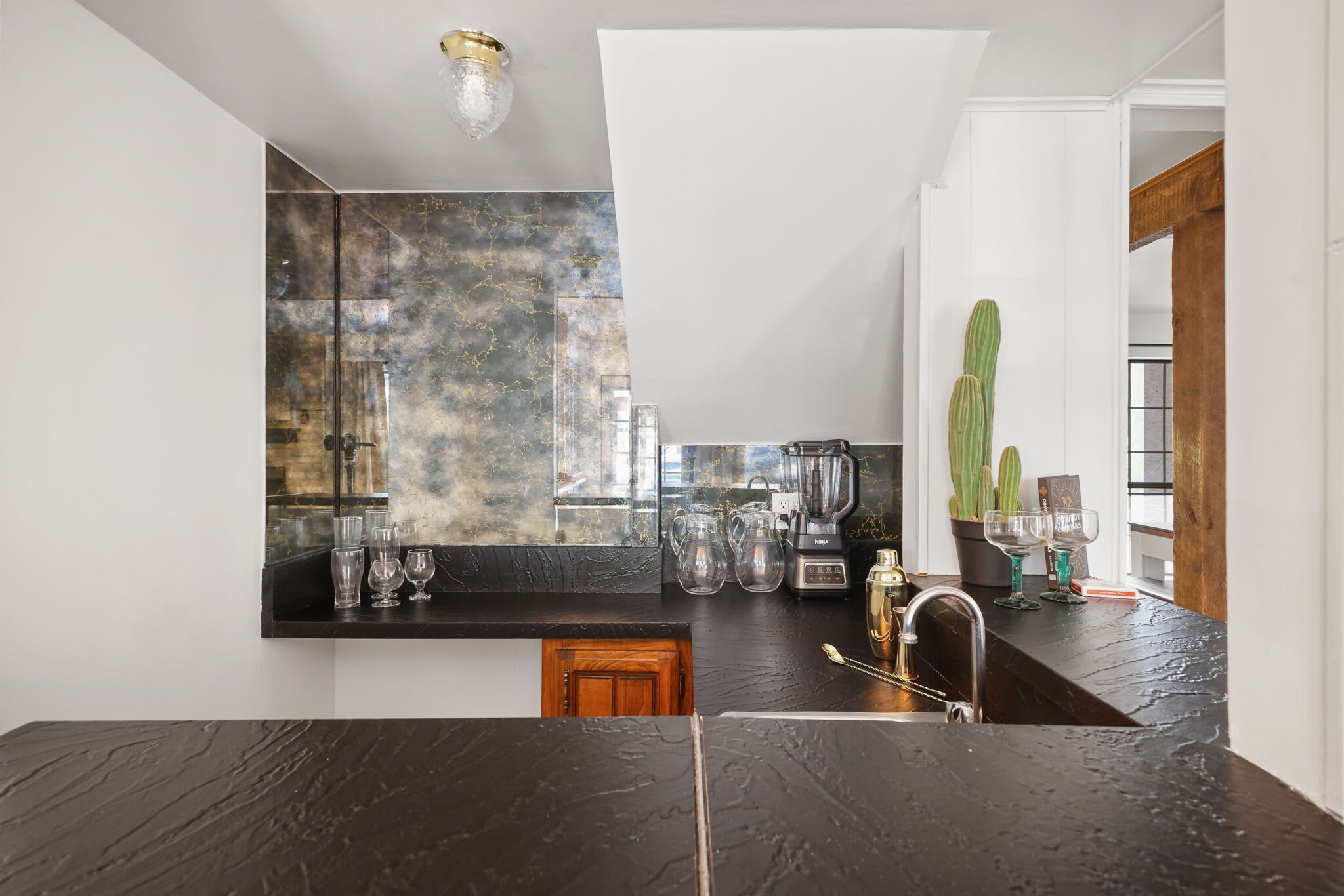 Una cocina moderna con encimera negra, salpicadero con diseño de mármol y un fregadero. Hay varios utensilios de cocina sobre la encimera, incluyendo una licuadora, una cafetera y cristalería.