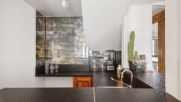 Una cocina moderna con encimera negra, salpicadero con diseño de mármol y un fregadero. Hay varios utensilios de cocina sobre la encimera, incluyendo una licuadora, una cafetera y cristalería.