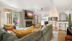 A modern living room with a fireplace, a sofa with colorful cushions, a large painting above the fireplace, and a neon sign in the background.