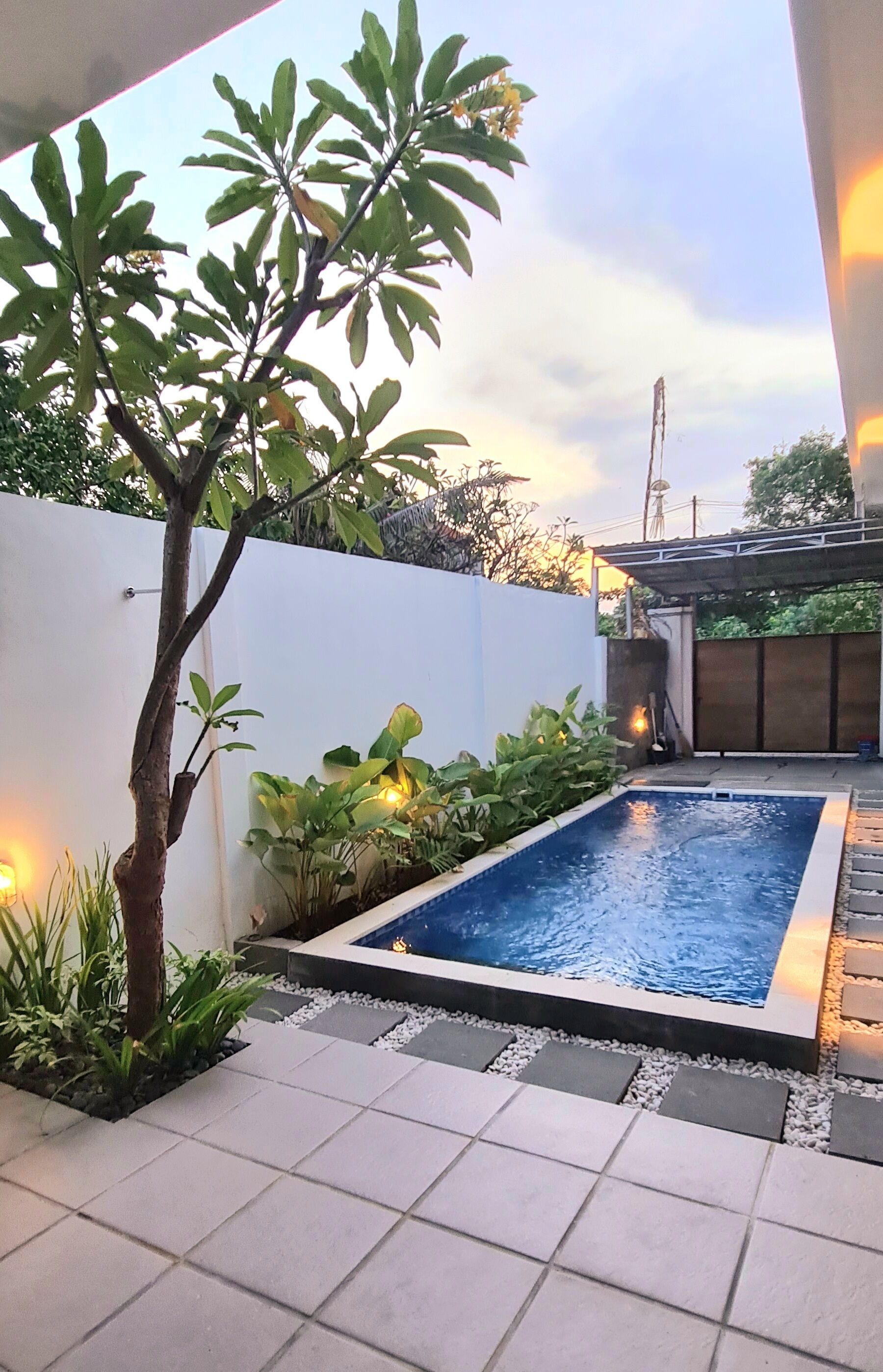 Un área de piscina exterior moderna con un deck de azulejos, rodeada de plantas y una pared blanca.