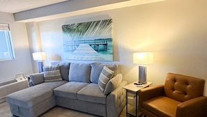 A living room with a sectional sofa, a brown armchair, a side table with a lamp, and a painting of a beach scene. - Sunset Hollow/Premium Location (Myrtle Beach)