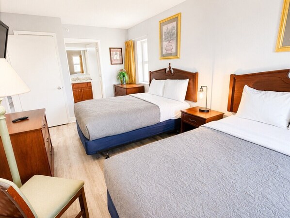 A hotel room with two beds, a desk, a chair, a bedside table, a lamp, and a window with curtains. - Wake up to the waves (Myrtle Beach)
