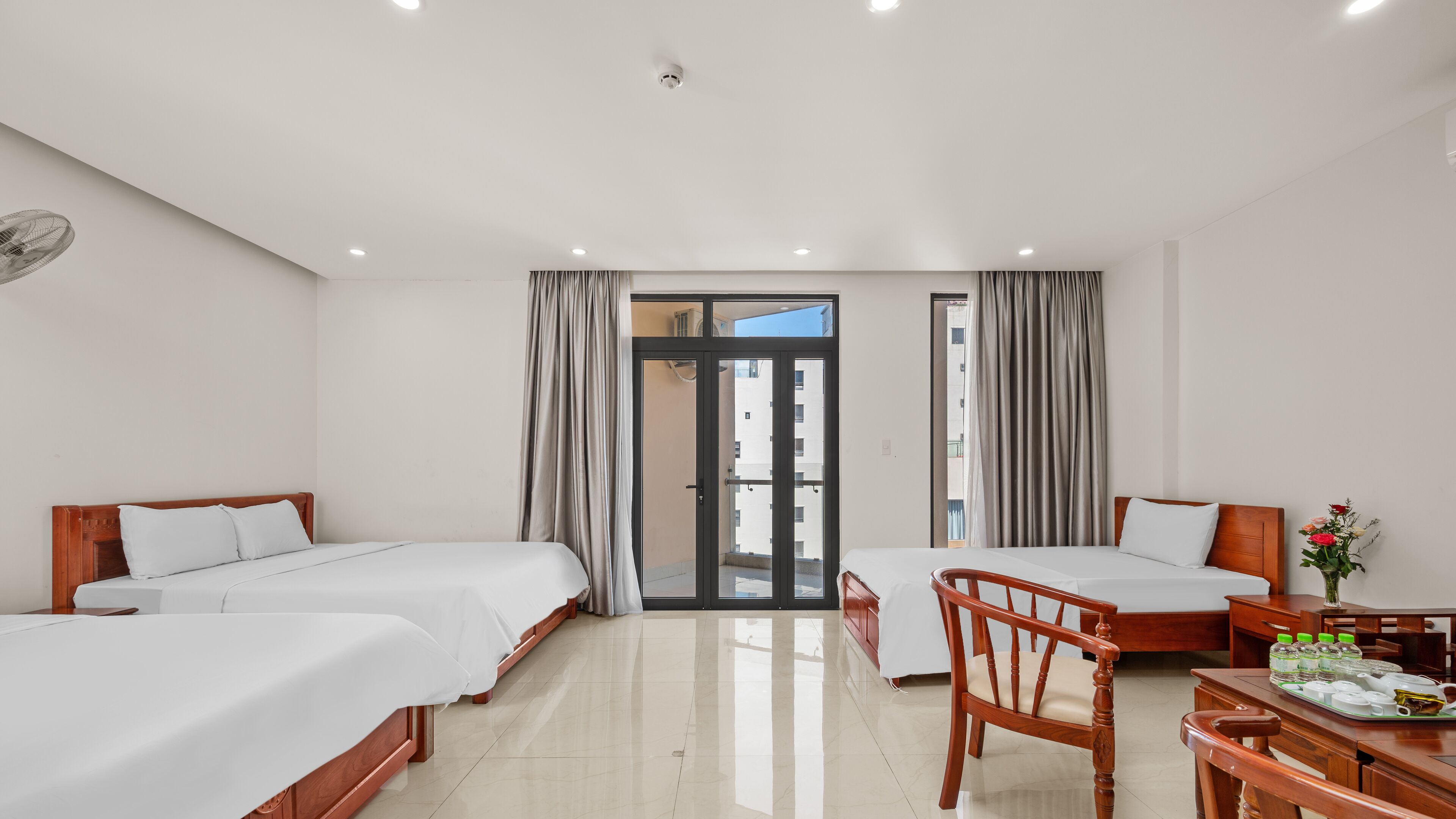 A hotel room with two beds, a wooden chair, a small table with a vase of flowers, and a view of a building through the door.