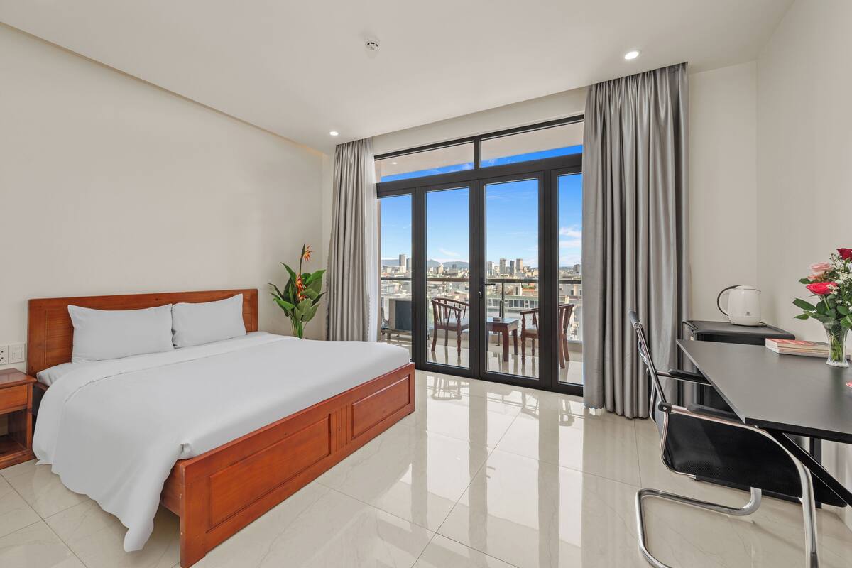 Una camera d'albergo moderna con un letto in legno, una scrivania e vista sulla città attraverso ampie finestre.