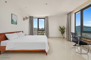 Habitación de hotel con una cama grande, un escritorio, una silla, una planta en maceta y vistas a la ciudad y al mar.