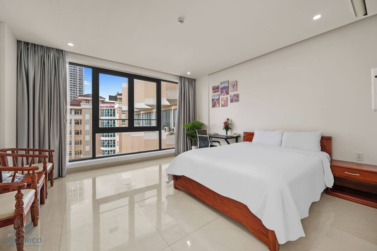 Camera d'albergo con un letto grande, mobili in legno, una scrivania e vista sugli edifici attraverso un'ampia finestra.