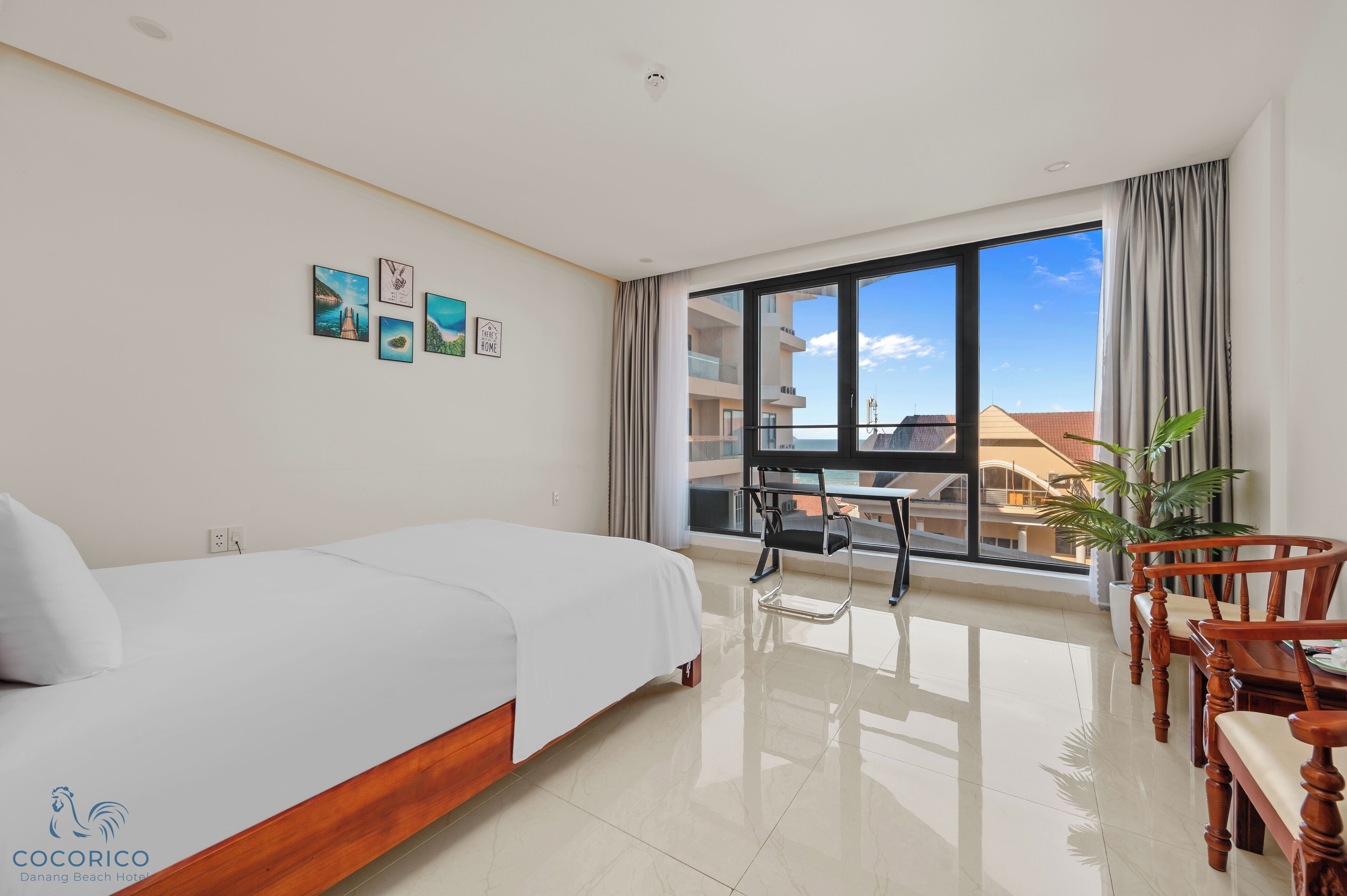 A hotel room with a bed, a dining table with chairs, a potted plant, and a large window offering a view of buildings and a clear sky.