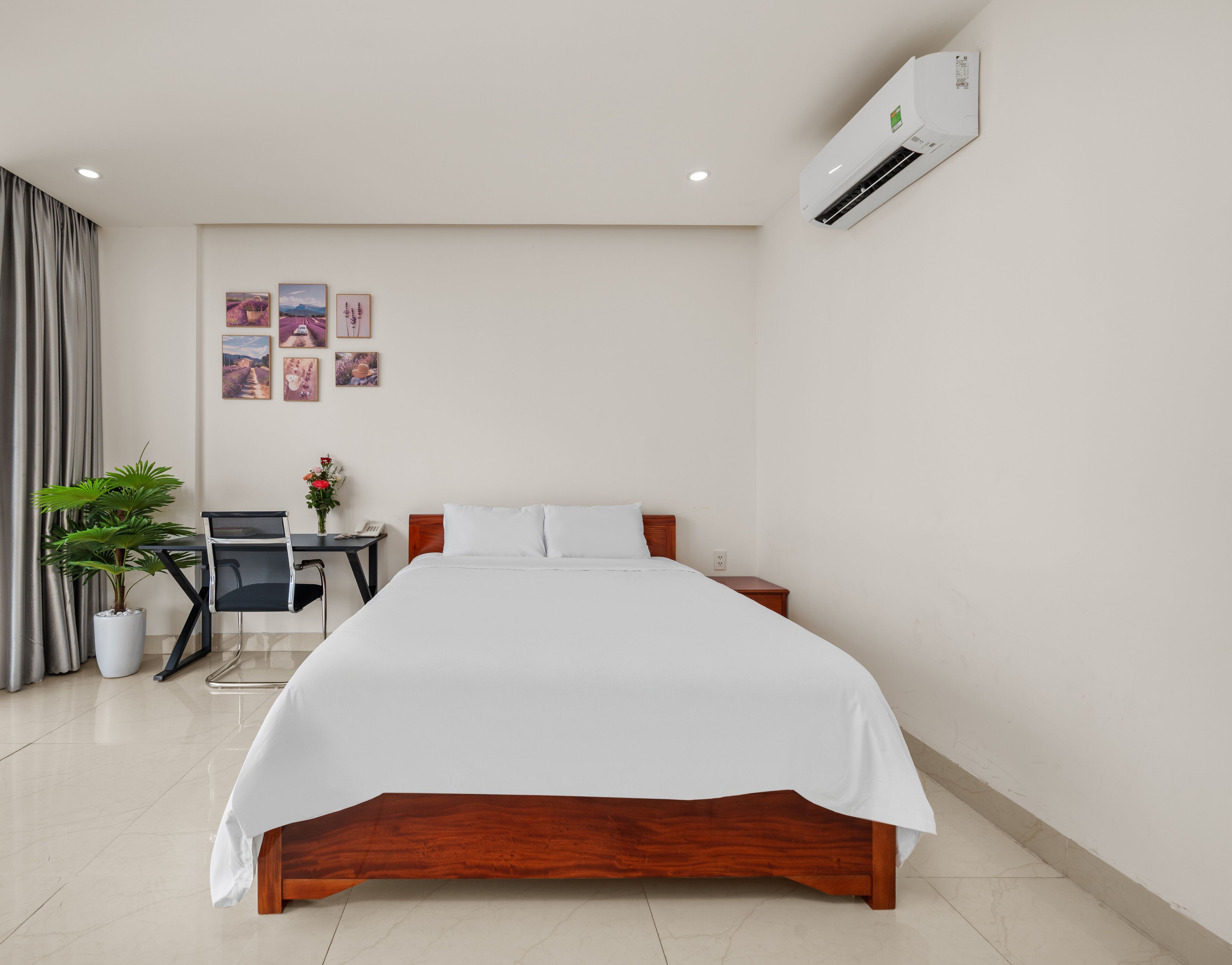 A hotel room with a wooden bed, a desk with a chair, a potted plant, and a wall-mounted air conditioner.