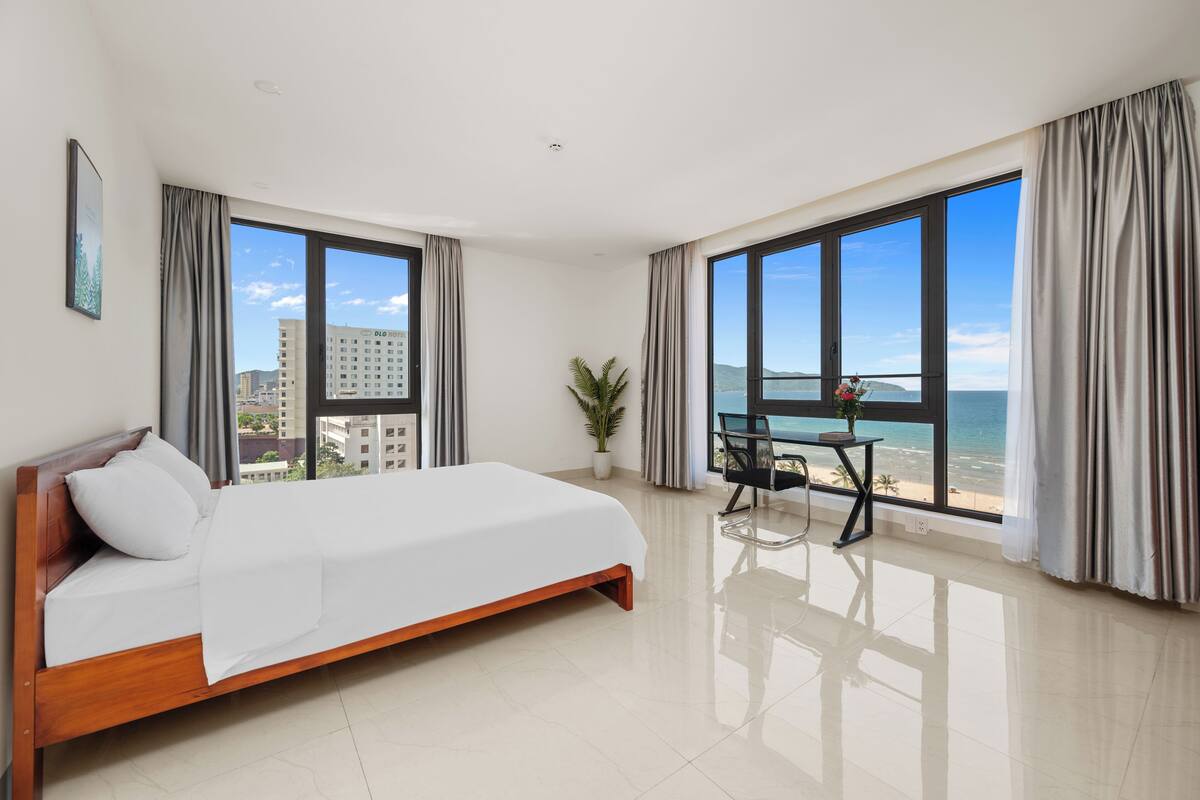 Una camera d'albergo moderna con un letto grande, un tavolino con una sedia e vista sulla spiaggia e sull'oceano attraverso ampie finestre.