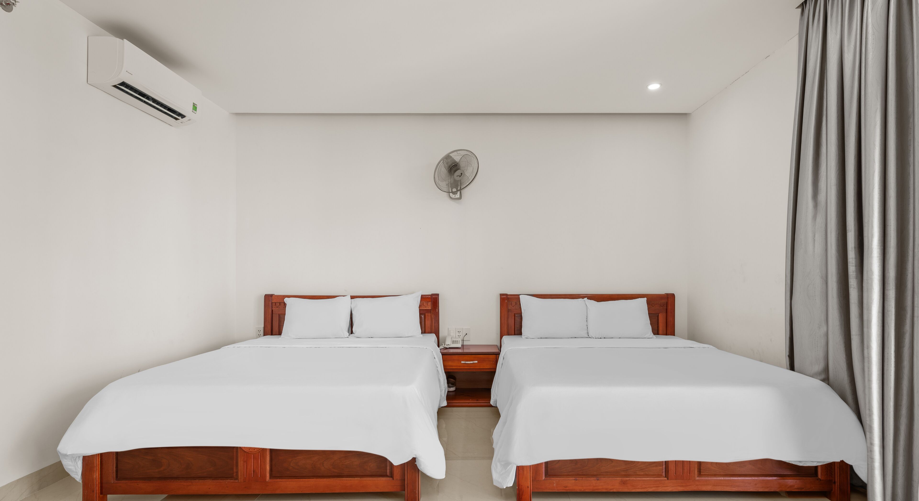 Una habitación de hotel con dos camas de madera, ropa blanca, un ventilador de pared y una ventana con cortinas.