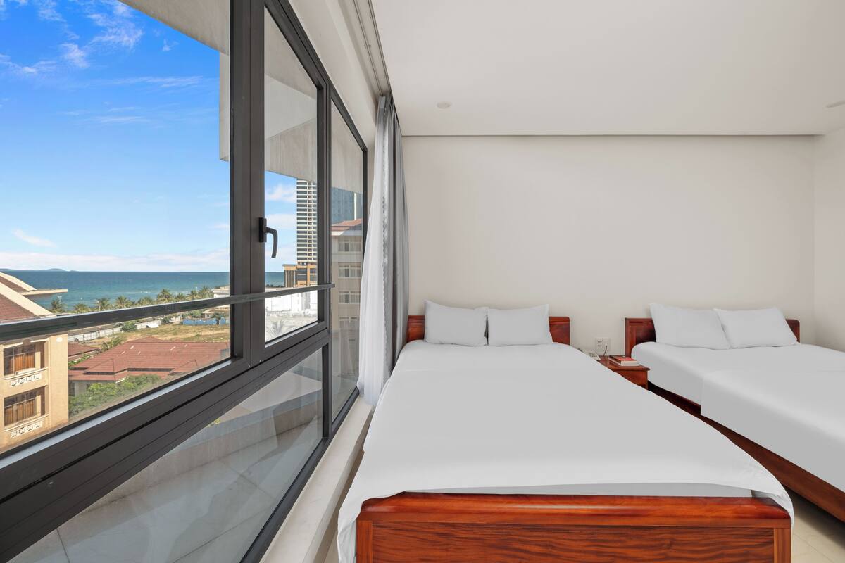 Camera d'albergo con un'ampia finestra che si affaccia sull'oceano e sugli edifici. Sono presenti due letti con lenzuola bianche e testiere in legno.