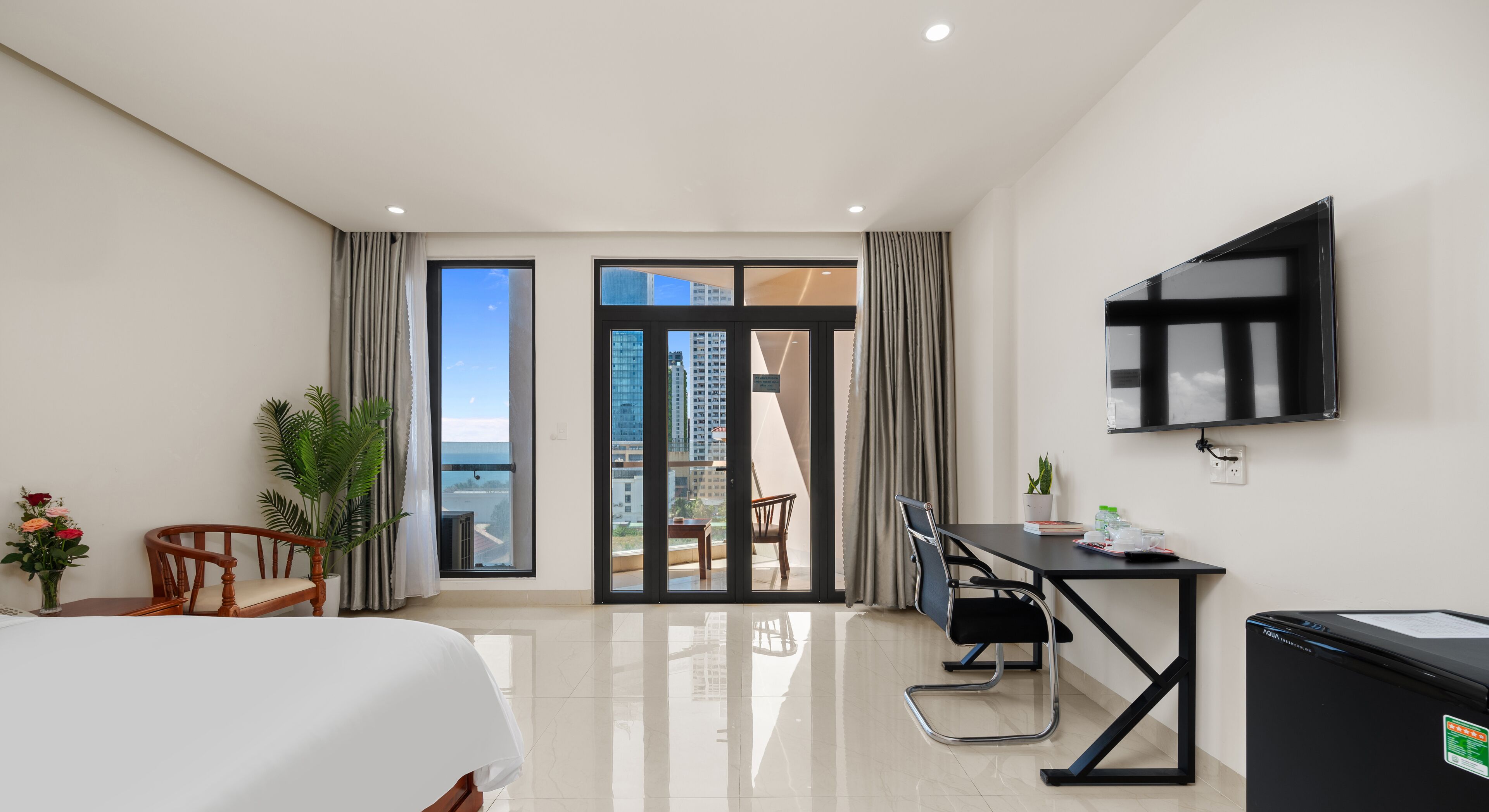 A hotel room with a bed, a desk, a chair, a TV, and a balcony with a view of buildings.