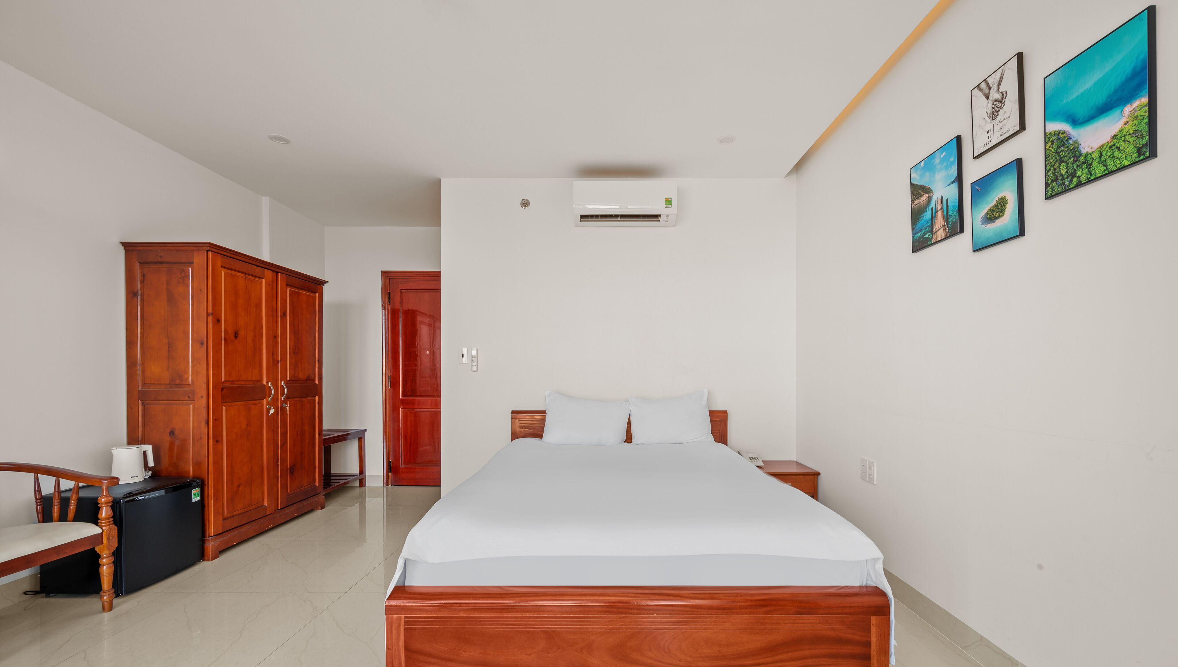 A bedroom with a bed, wooden wardrobe, a small table with a kettle, and a refrigerator.