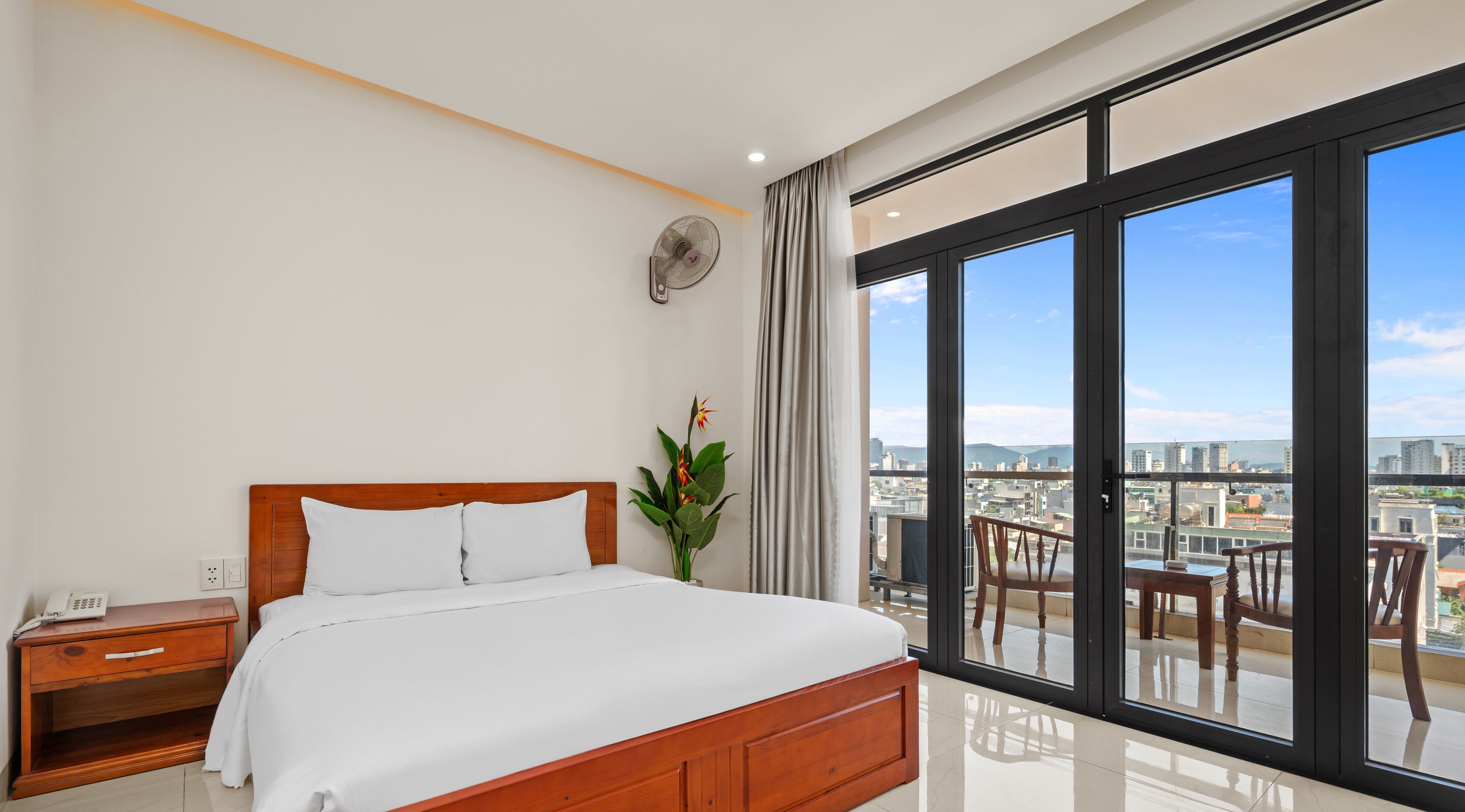 A modern hotel room with a large bed, a bedside table with a phone, a plant, and a view of the city through large windows.