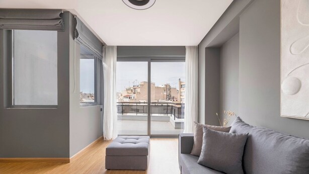En moderne stue med en grå sofa, en pudeskammel og et sofabord. Der er et stort vindue med udsigt til bygninger og en balkon.