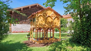 Una casita de juegos de madera con techo inclinado y un pequeño porche, rodeada de un césped cuidado y un muro de piedra.