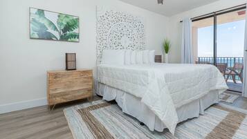 Un dormitorio con cama, una mesita de noche de madera, un panel decorativo en la pared, una planta y vista al océano desde un balcón.
