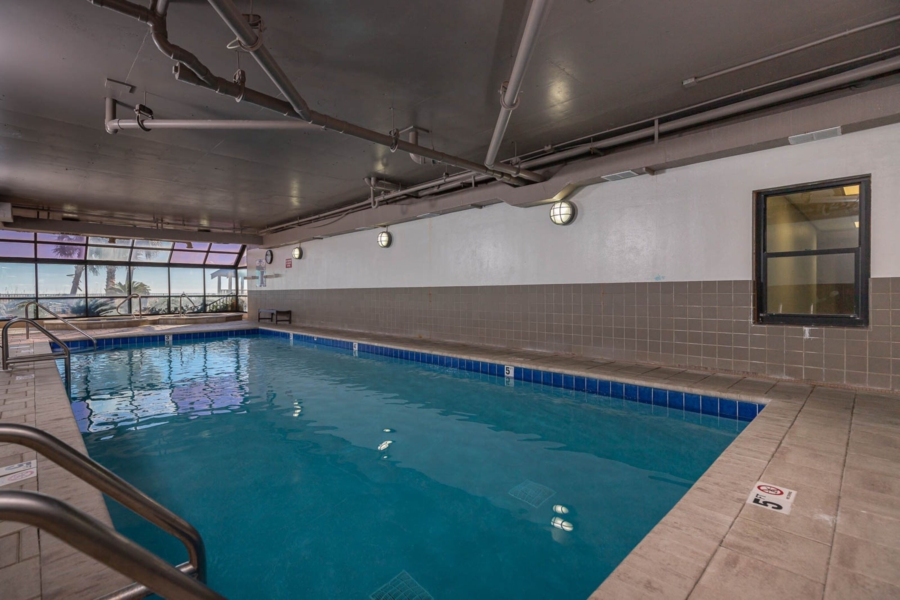 An indoor swimming pool with clear water, tiled walls, and a window providing a view of palm trees.
