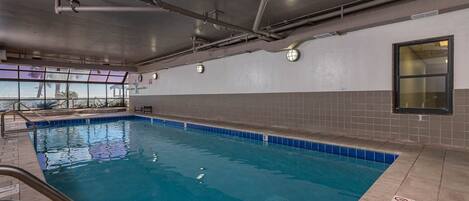 An indoor swimming pool with clear water, tiled walls, and a window providing a view of palm trees.