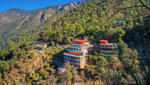 Un hotel de varios pisos con balcones, situado en una zona montañosa con un bosque denso.