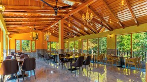 Un restaurante espacioso con techos altos, vigas de madera y amplias ventanas que ofrecen vistas a zonas verdes. Mesas y sillas dispuestas para comer.