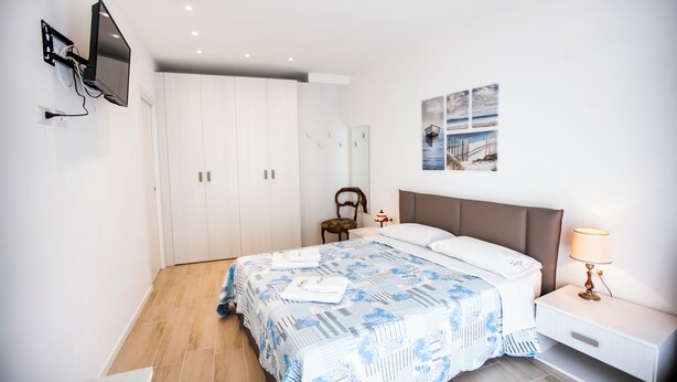 A bedroom with a bed, a nightstand, a lamp, a TV mounted on the wall, and a wardrobe. - Benzi16 (Como)