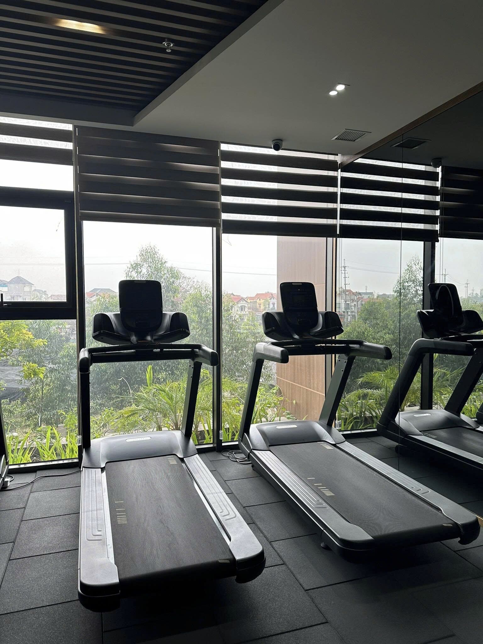 A gym with three treadmills aligned in a row, large windows with blinds, and a view of greenery outside.