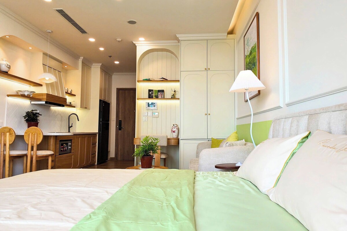 A hotel room with a bed, a desk, a chair, a small table, a lamp, a wardrobe, and a potted plant.